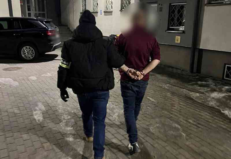 Policjant prowadzi zatrzymanego mężczyznę z założonymi kajdankami na podwórzu nocą; twarz zatrzymanego jest zamazana.